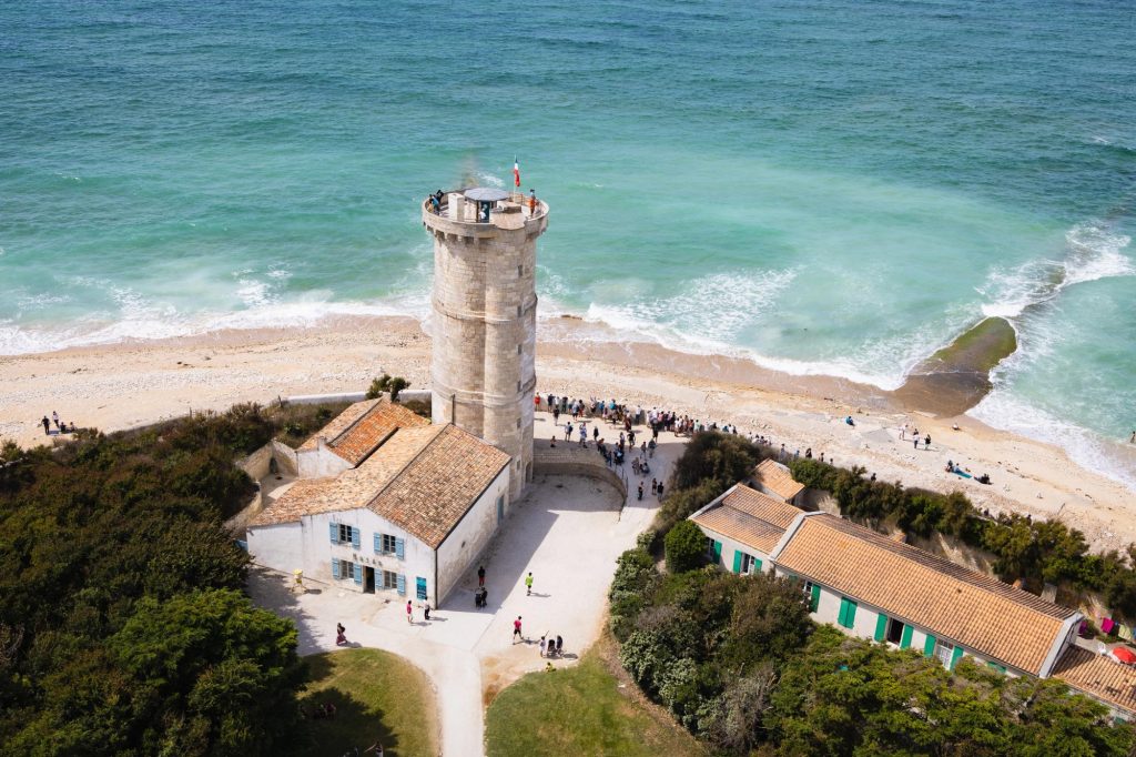 Aerial view of a lighthouse by the beach with people gathered around, turquoise ocean waves and sandy shore in the background.
