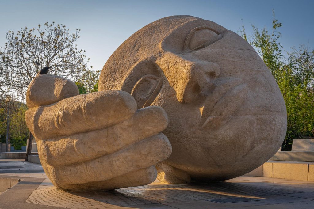The Giant Head at Les Halles: a large stone sculpture depicting a human face and hand, symbolizing listening and contemplation, surrounded by greenery.