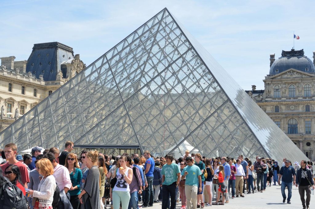 A long queue of visitors waiting to enter the Louvre Museum, showcasing the iconic glass pyramid in the foreground and the historic architecture in the background.