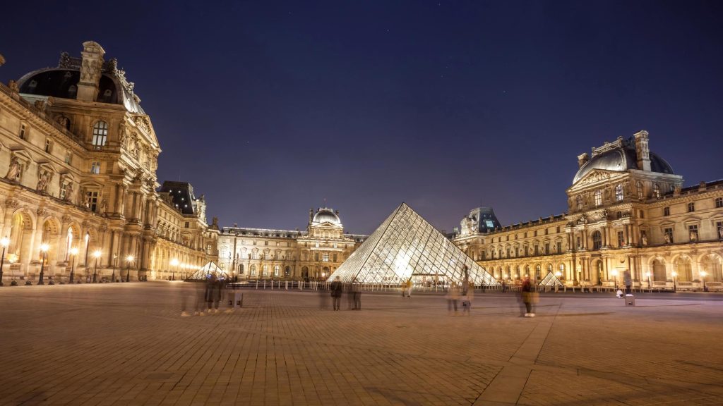 A nighttime view of the Louvre Museum with the glass pyramid prominently displayed, illuminated by lights, and visitors in motion around the plaza.