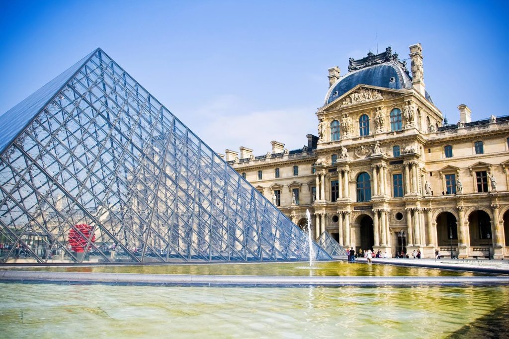View of the glass pyramid of the Louvre Museum with the historic building in the background under a blue sky.