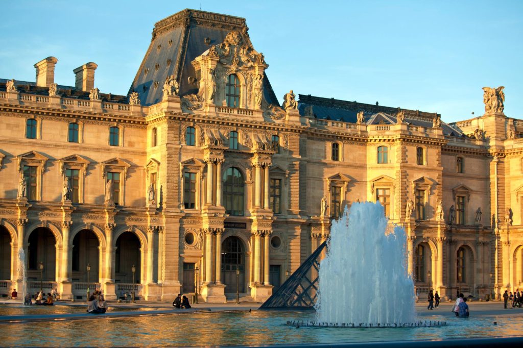 View of the Louvre Palace and pyramid during sunset with a fountain in the foreground.