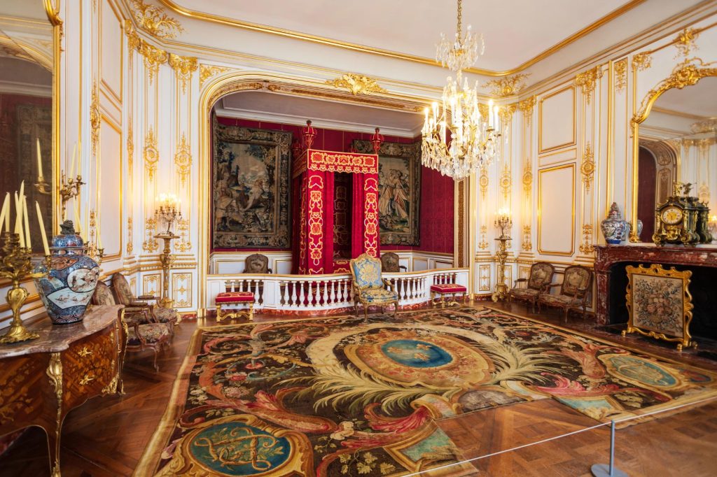 Interior of a luxurious room featuring ornate golden decorations, elegant furniture, and a large chandelier, depicting rich historical aesthetics.