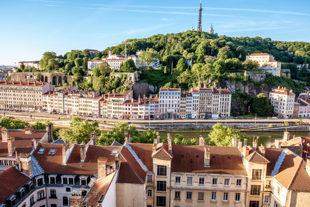 A picturesque view of the city of Lyon, France, showcasing colorful buildings along a river with a hilltop covered in greenery and historical structures.