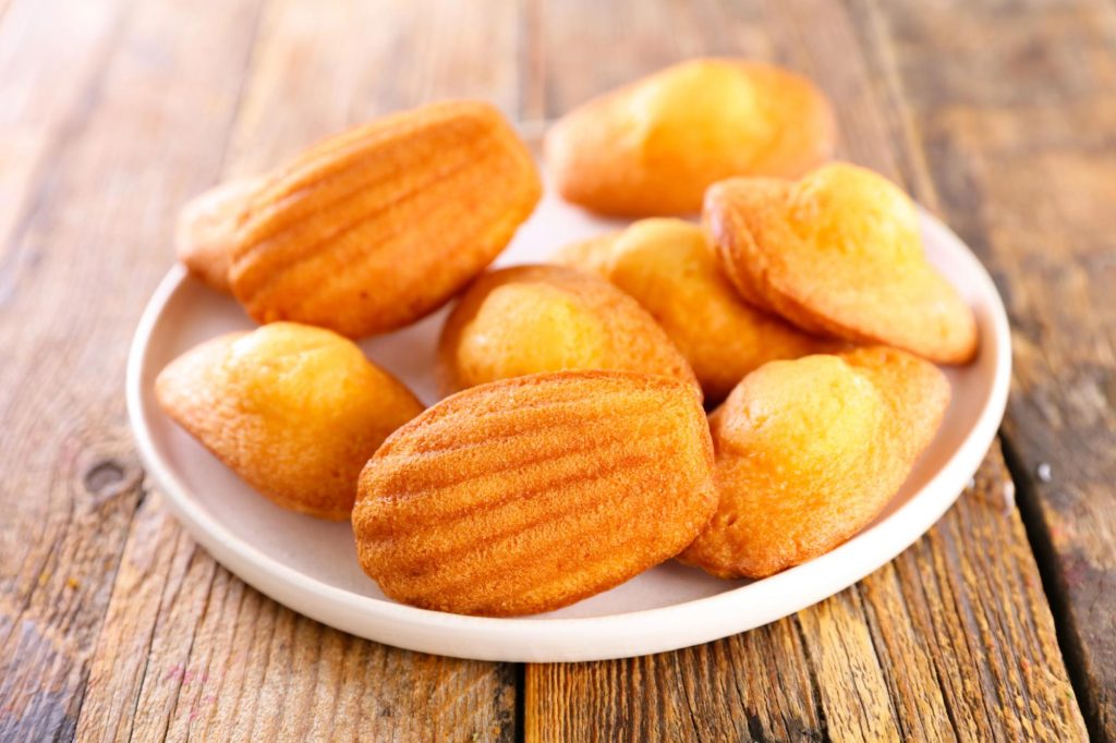 A plate of golden-brown madeleines, French sponge cakes with a distinctive shell shape, sitting on a rustic wooden surface.