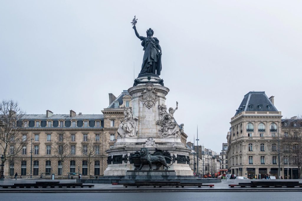 A statue of Marianne, symbolizing the French Republic, stands prominently in Place de la République, surrounded by historical buildings and a cloudy sky.