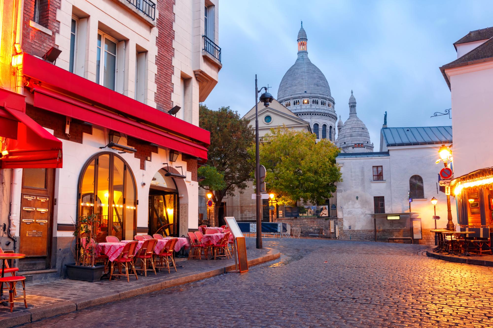 A charming street in Montmartre, Paris, featuring a café with outdoor seating and a view of the Basilica of Sacré-Cœur under a twilight sky.