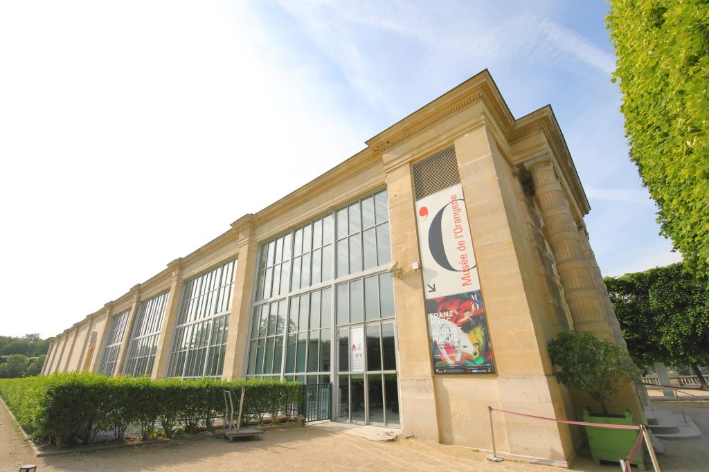 Exterior view of the Musée de l'Orangerie in Paris, featuring large glass windows and a promotional banner, surrounded by lush greenery.