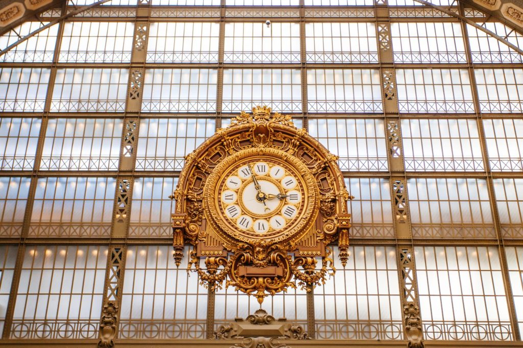 A beautiful ornate clock on display in a museum, featuring Roman numerals and intricate designs, set against a backdrop of large windows.