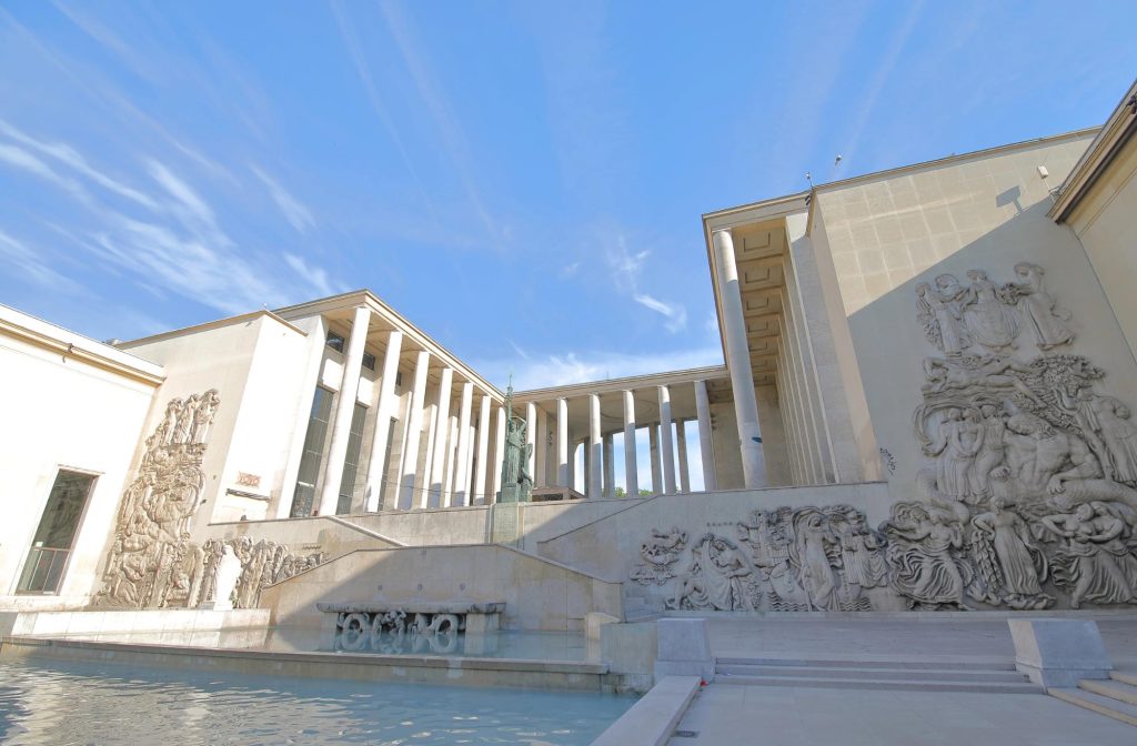 Exterior view of the Museum of Modern Art in Paris featuring a fountain and sculptures on the building's facade under a clear blue sky.