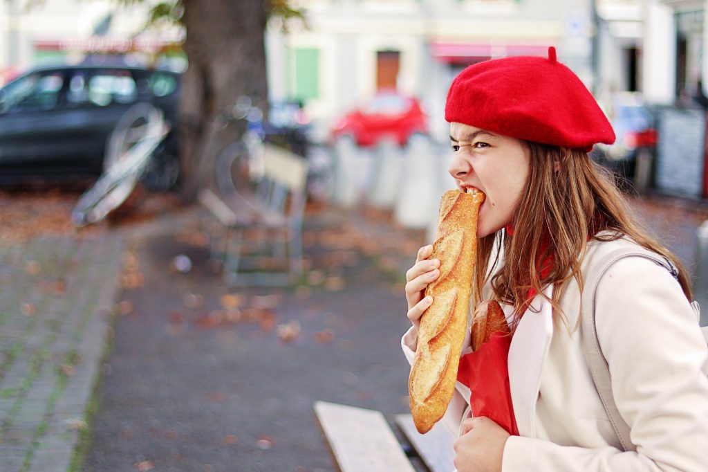 A young woman in a red beret biting into a fresh baguette, sitting in a park with autumn leaves on the ground.