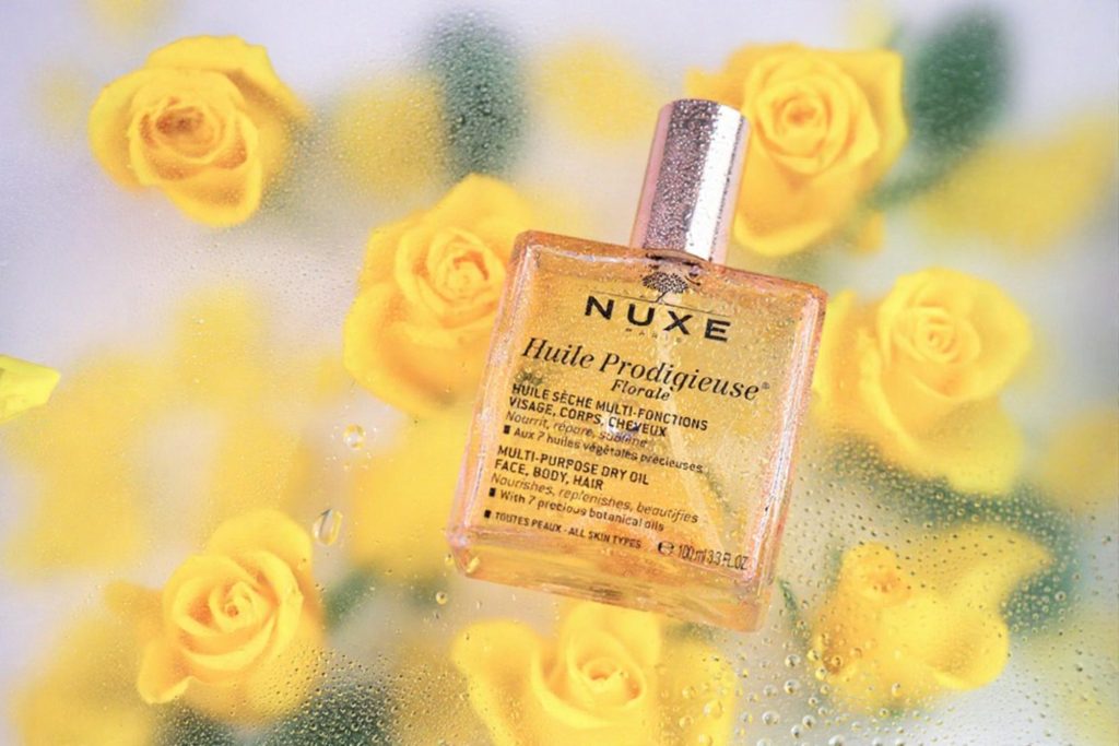 A bottle of Nuxe Huile Prodigieuse dry oil surrounded by yellow roses and droplets of water.