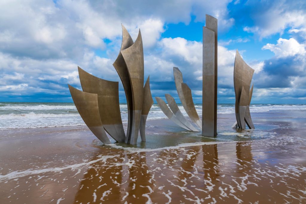 Sculpture commemorating the Omaha Beach landing, with abstract metallic shapes rising from the sandy shore, under a cloudy sky.