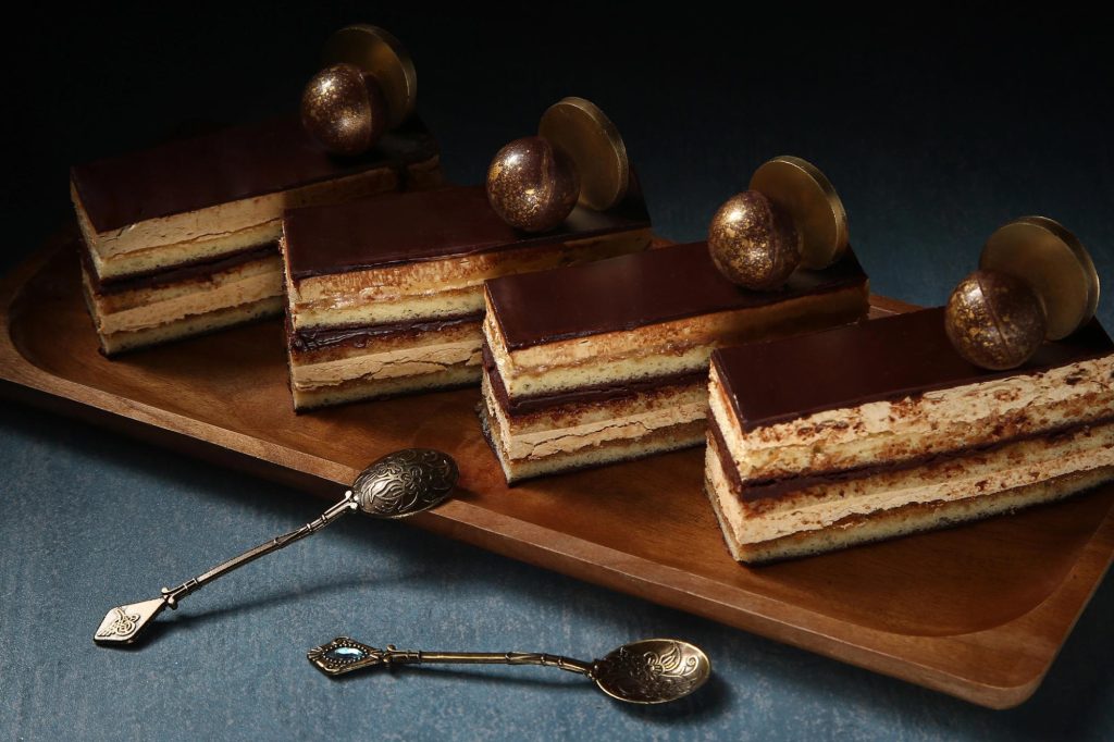 A close-up of slices of opera cake displayed on a wooden platter, featuring layers of chocolate, coffee, and almond sponge with decorative spherical toppings and ornate silver spoons nearby.