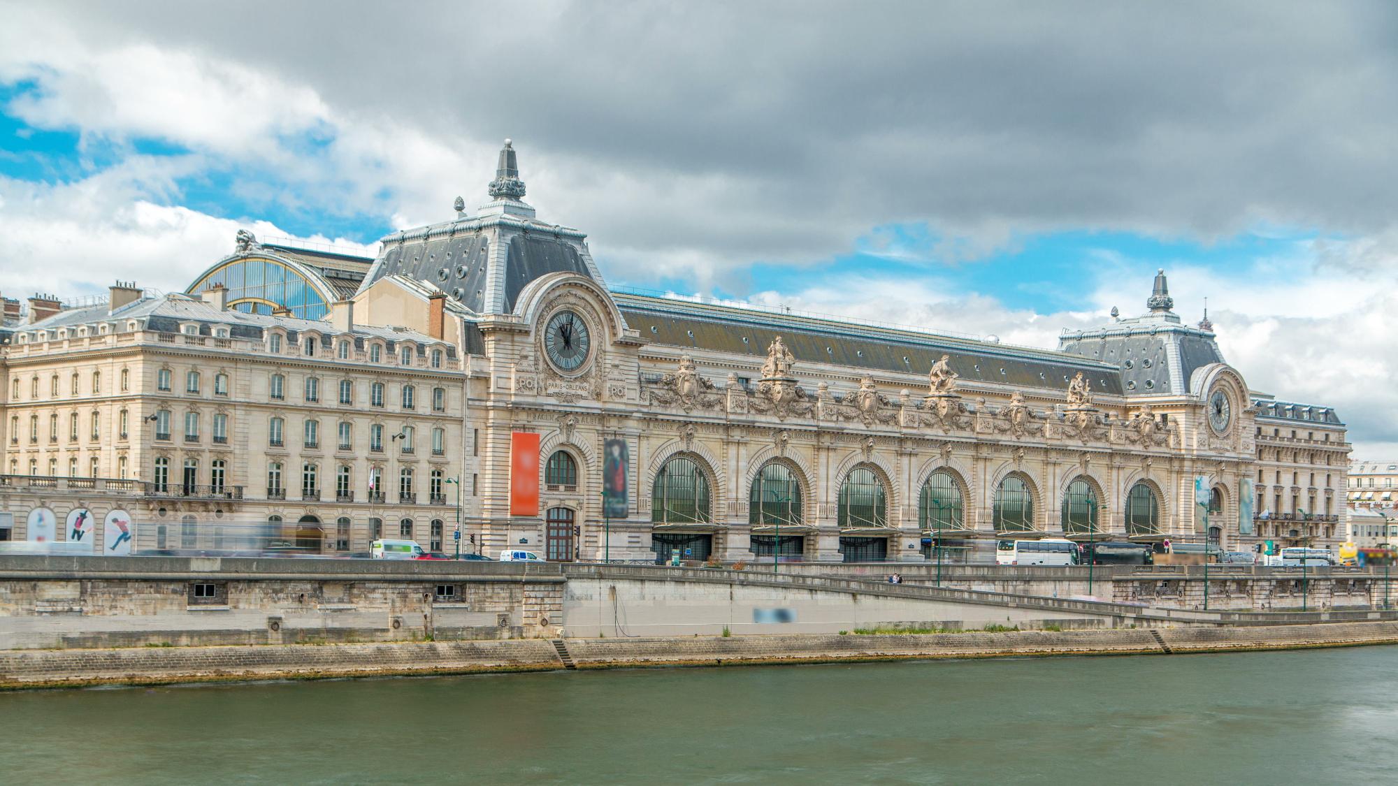 A view of the Musée d'Orsay building in Paris, showcasing its ornate architecture and clock tower along the Seine River under a cloudy sky.