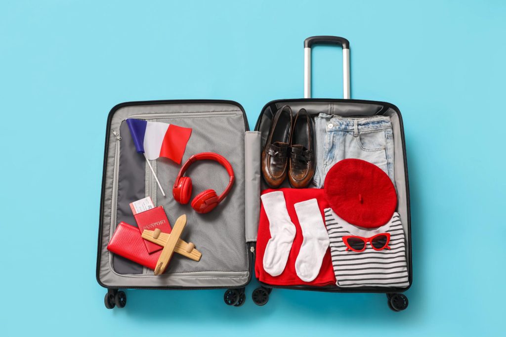 A suitcase packed for a trip to France, featuring a French flag, passport, red headphones, shoes, a red beret, striped shirt, and rolled-up clothing items.
