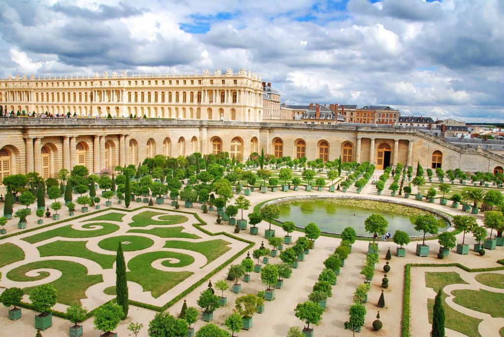 A beautiful view of the gardens at the Palace of Versailles, showcasing the intricate landscaping, neatly arranged trees, and decorative water features under a cloudy sky.