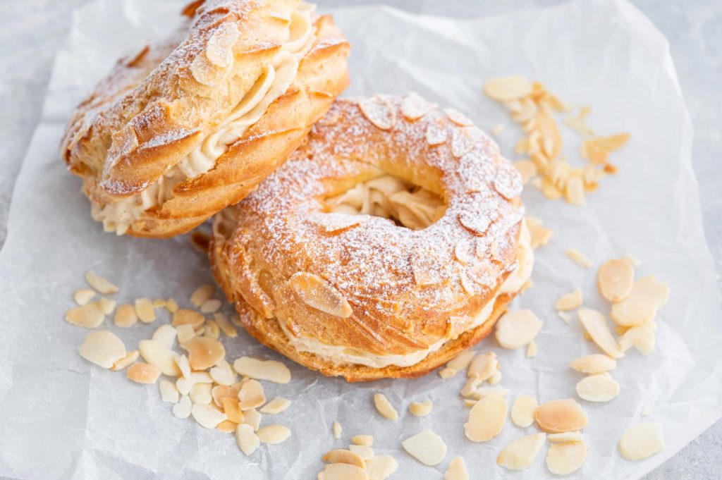 Two delicious Paris-Brest pastries made of choux pastry filled with cream, sprinkled with powdered sugar and almond slivers on parchment paper.