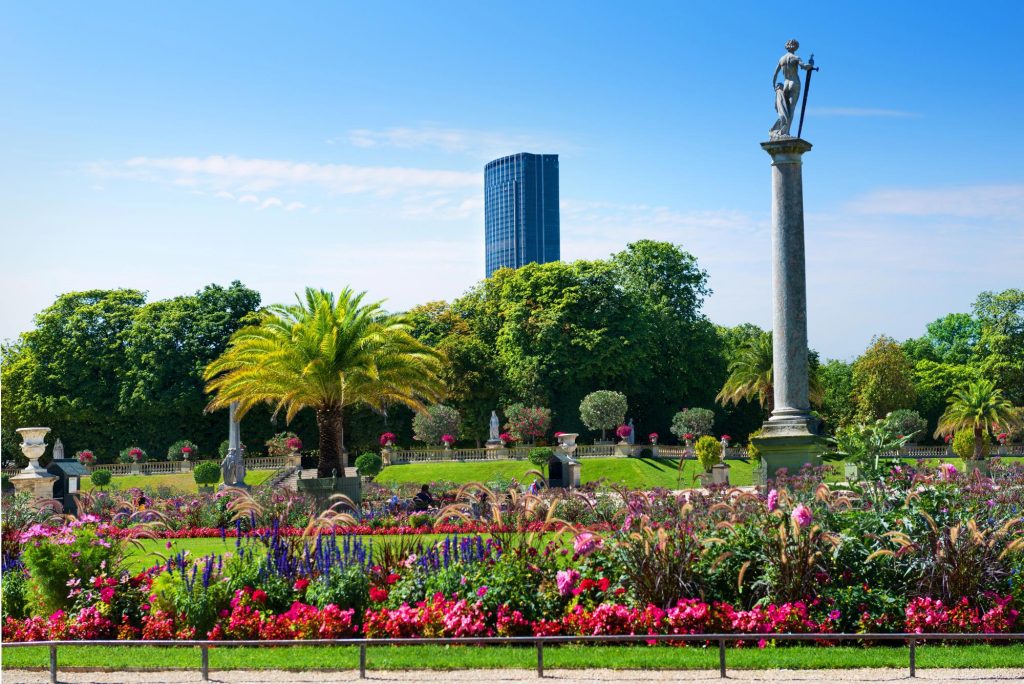 A beautiful park with colorful flowers and lush greenery, featuring a tall statue on a column and a modern building in the background.