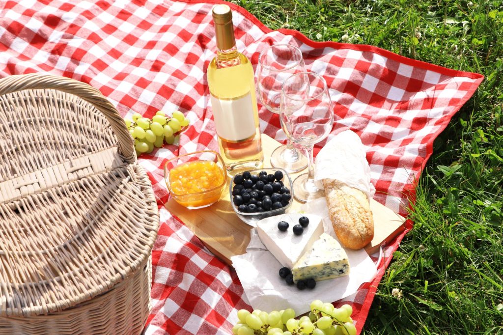 A picnic setup featuring a red and white checkered blanket, a wicker basket, glasses, a bottle of wine, and an assortment of delicious food including bread, cheese, blueberries, and grapes.
