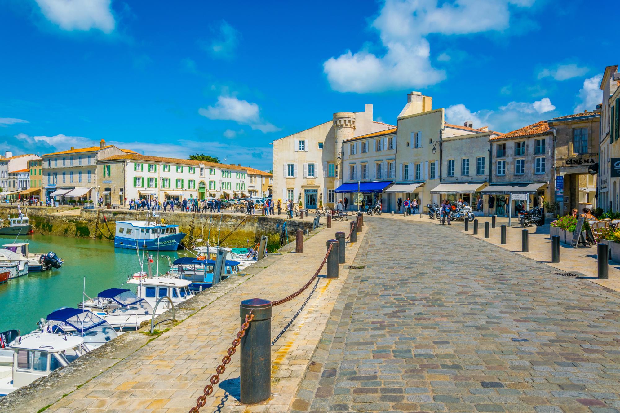 A lively harbor scene featuring boats, a cobblestone path, and charming buildings under a bright blue sky.