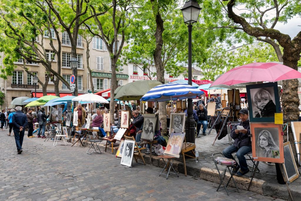 Artists displaying their artworks in Place du Tertre, Paris, with tourists walking by under umbrellas.