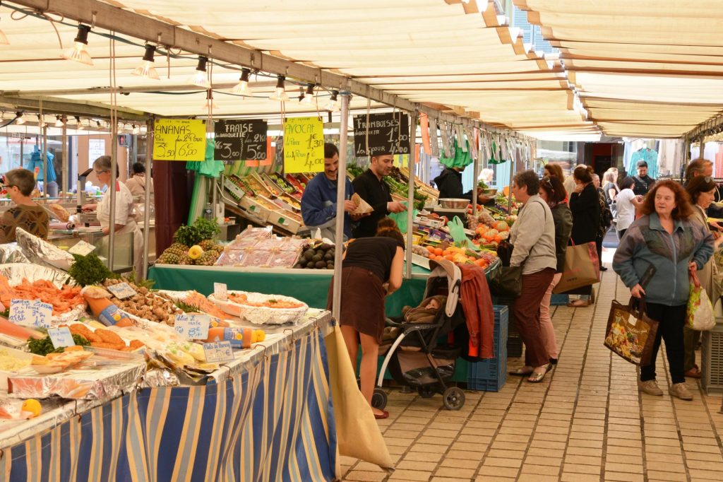 A bustling market scene with vendors selling fresh produce, seafood, and other goods under a canopy. Shoppers browse among stalls filled with colorful fruits and vegetables.