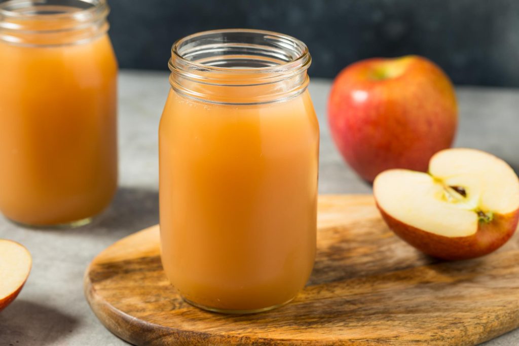 Two jars of cold apple cider juice on a wooden board with fresh apples, one whole and one halved.