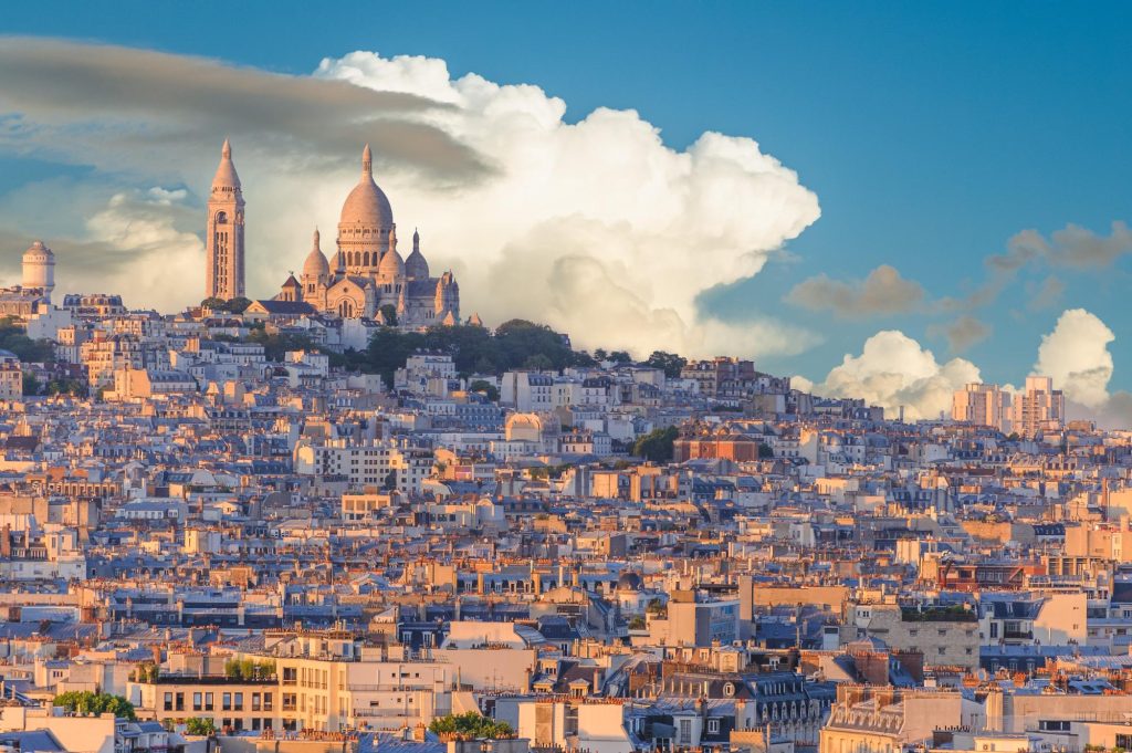 View of Sacré-Cœur Basilica on top of Montmartre hill in Paris, featuring a skyline of buildings and a cloudy sky.