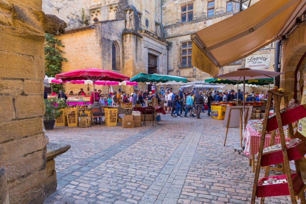 A lively market scene in Sarlat-la-Canéda featuring colorful stalls, visitors exploring local products, and historic stone buildings in the background.
