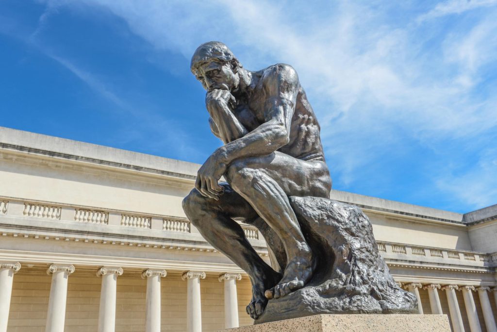 The Thinker, a bronze sculpture depicting a seated man deep in thought, with a clear blue sky and classical architecture in the background.