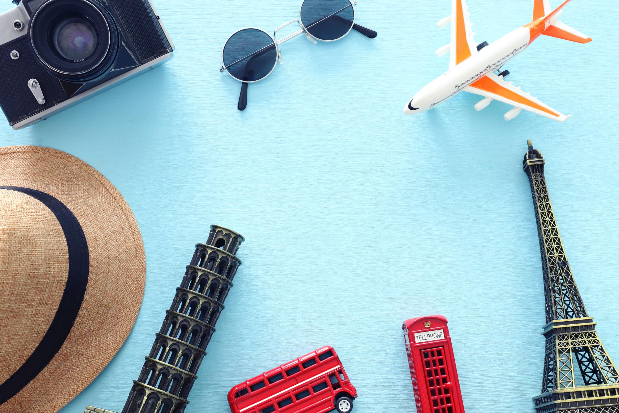 Flat lay of travel accessories including a vintage camera, sunglasses, a straw hat, miniature landmarks like the Eiffel Tower and the Leaning Tower of Pisa, a toy airplane, and a red double-decker bus.