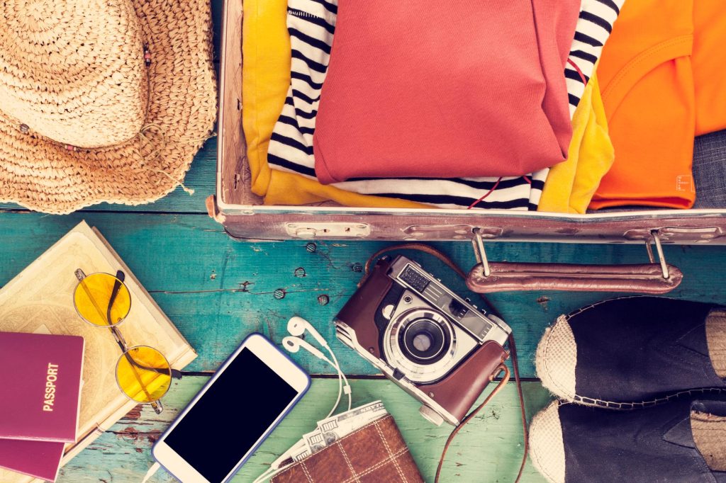 A flat lay of travel essentials including a suitcase filled with clothes, a straw hat, a vintage camera, a smartphone, sunglasses, a passport, and a wallet on a wooden table.