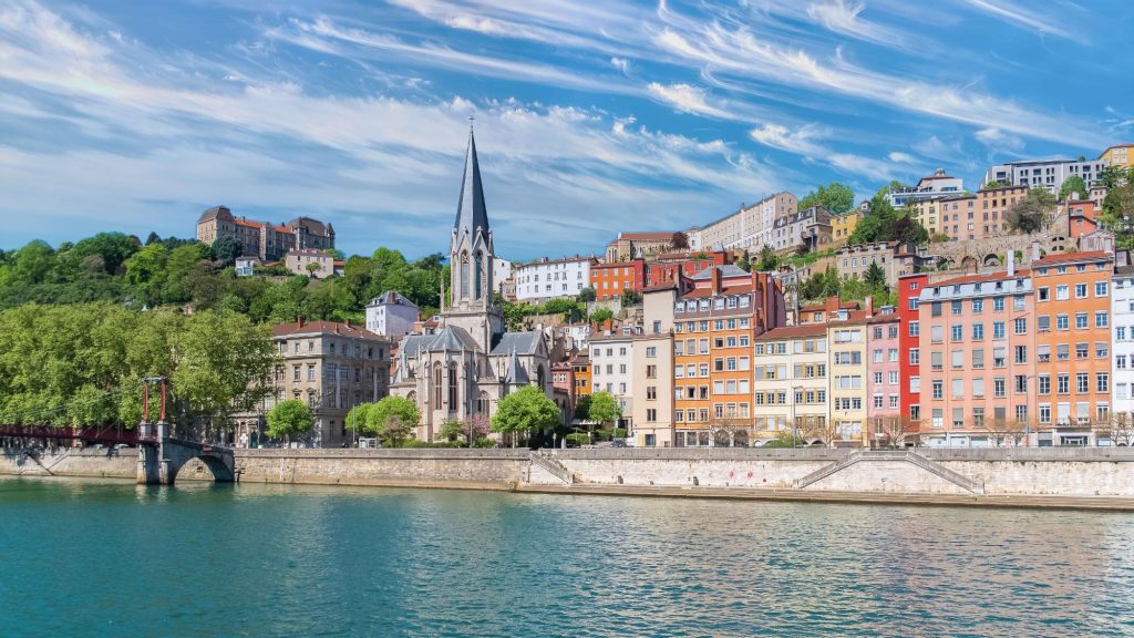 A scenic view of Saint Georges Church in Vieux Lyon with colorful houses along the riverbank under a bright blue sky.