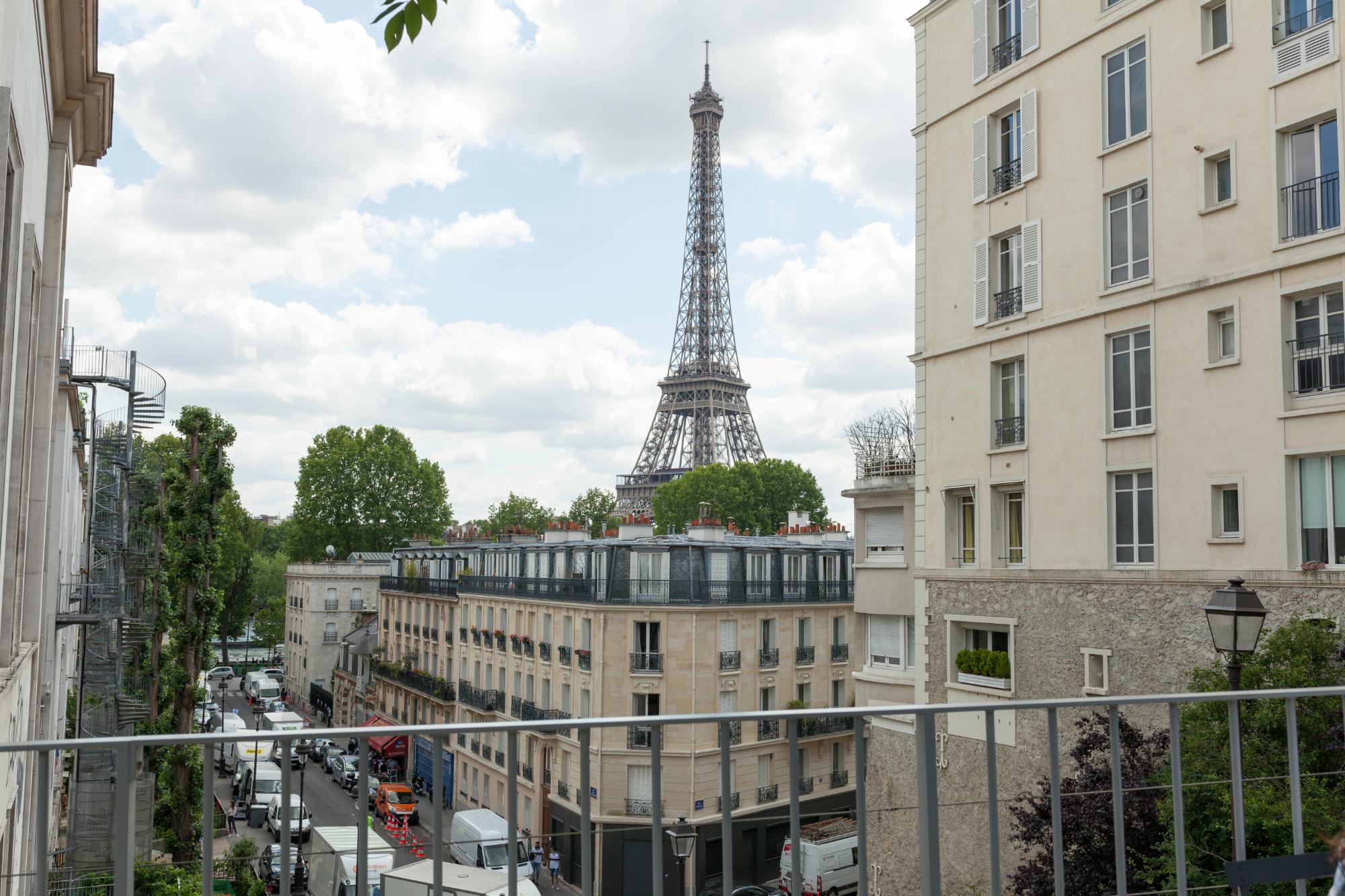 A scenic view of the Eiffel Tower towering over typical Parisian architecture, featuring trees and a bustling street below.