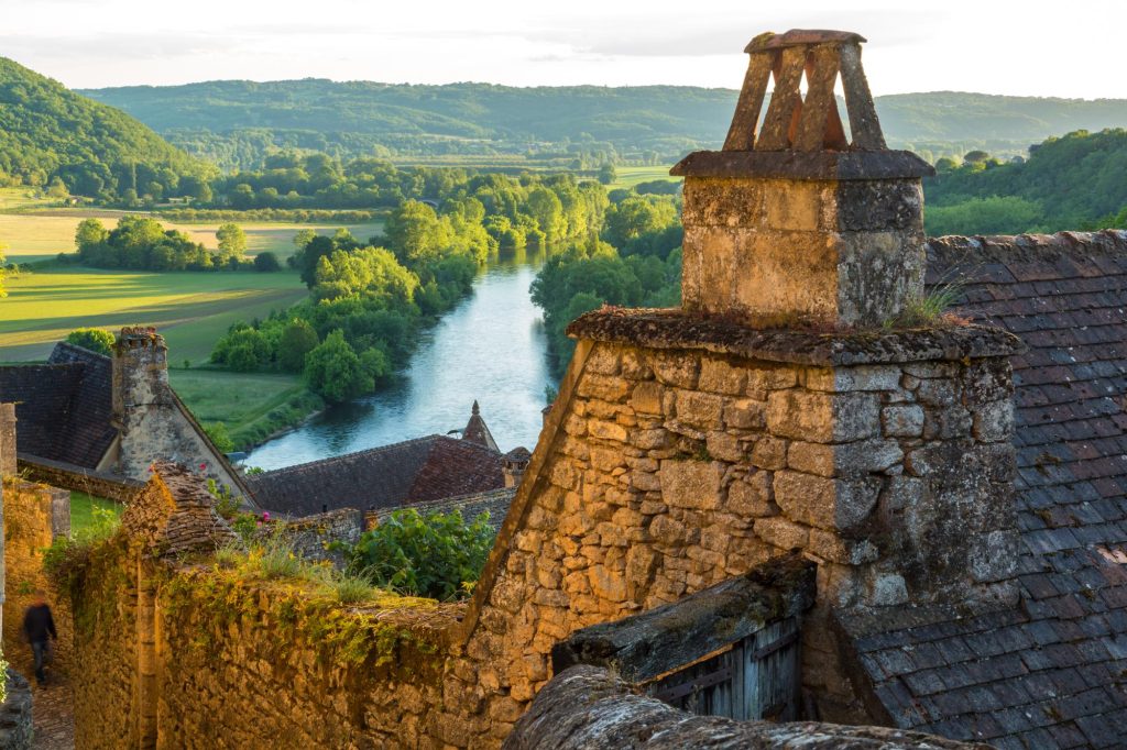 A scenic view of Beynac-et-Cazenac with the Dordogne River flowing through lush greenery and historic stone rooftops in the foreground.