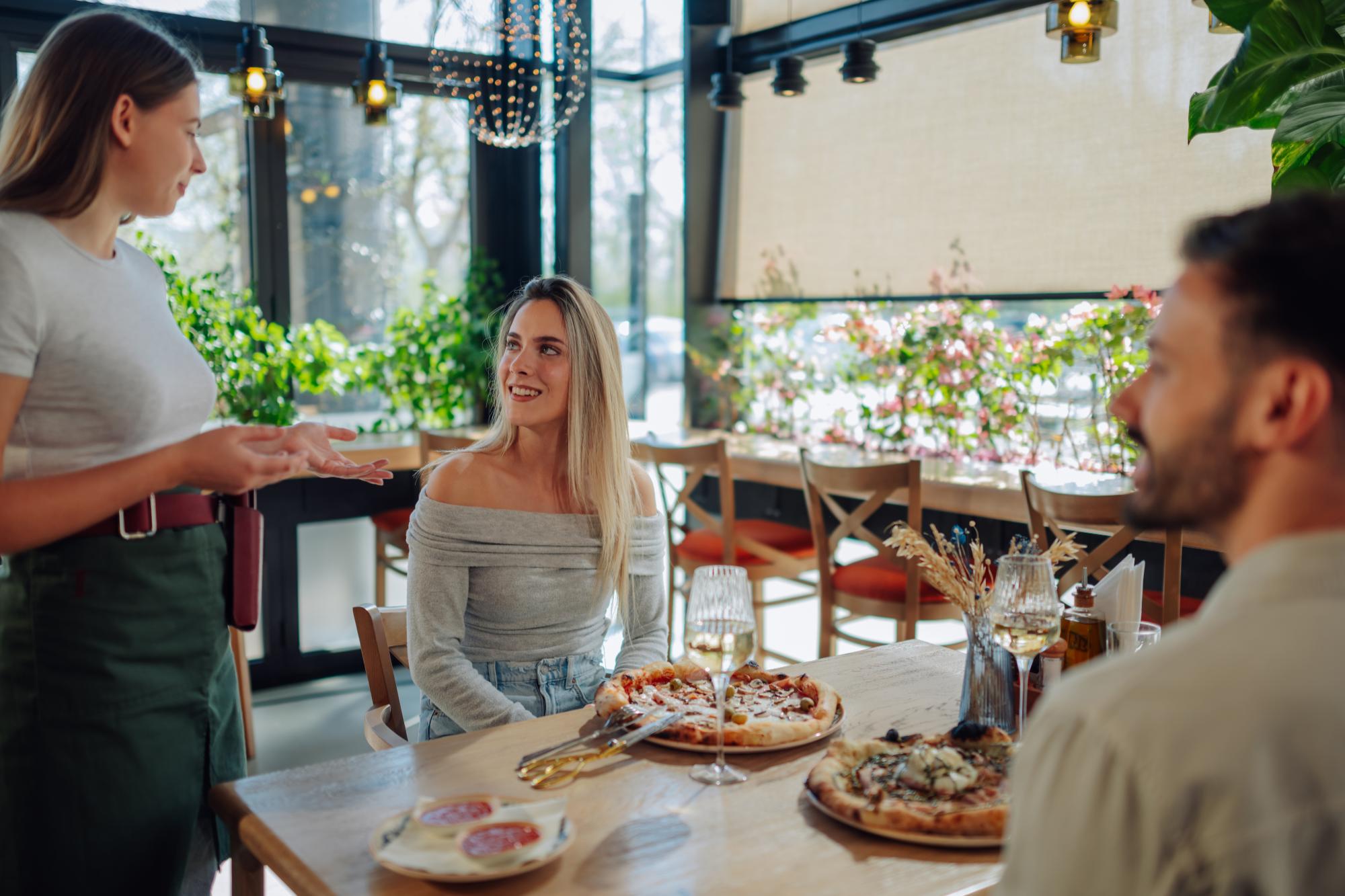 A waitress suggesting pizza to a couple dining in a modern restaurant with greenery and large windows.