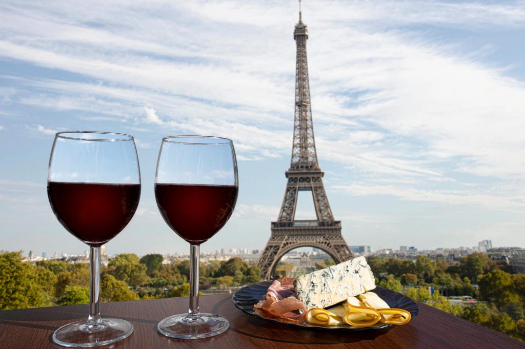 Two glasses of red wine beside a plate of cheese and cured meats, with the Eiffel Tower in the background against a blue sky.