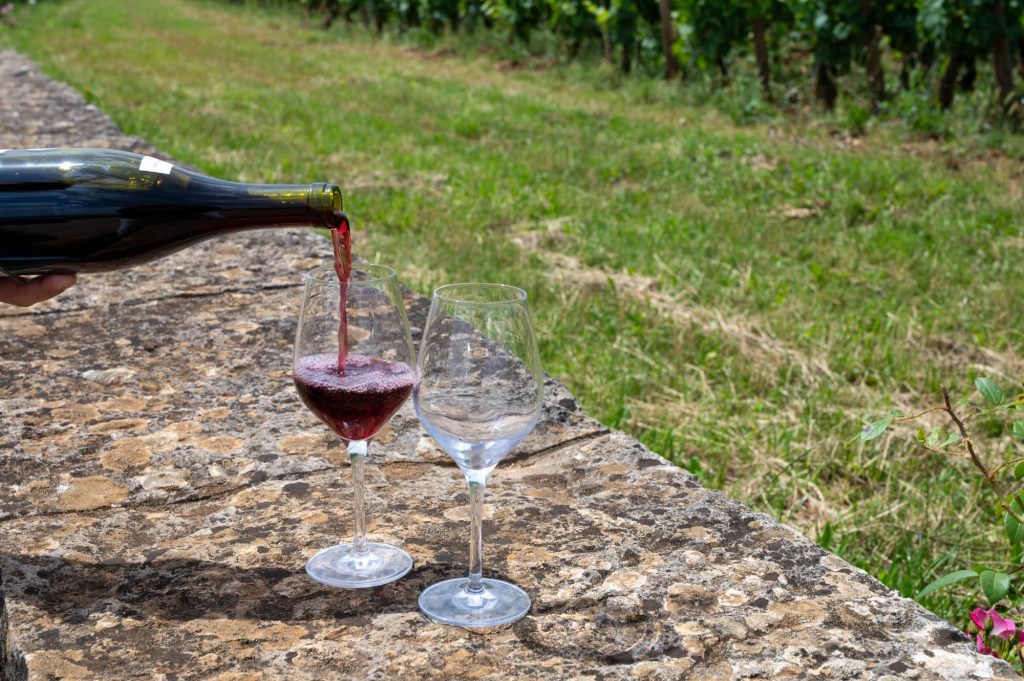 A bottle of red wine being poured into a glass on a stone surface with a vineyard background.