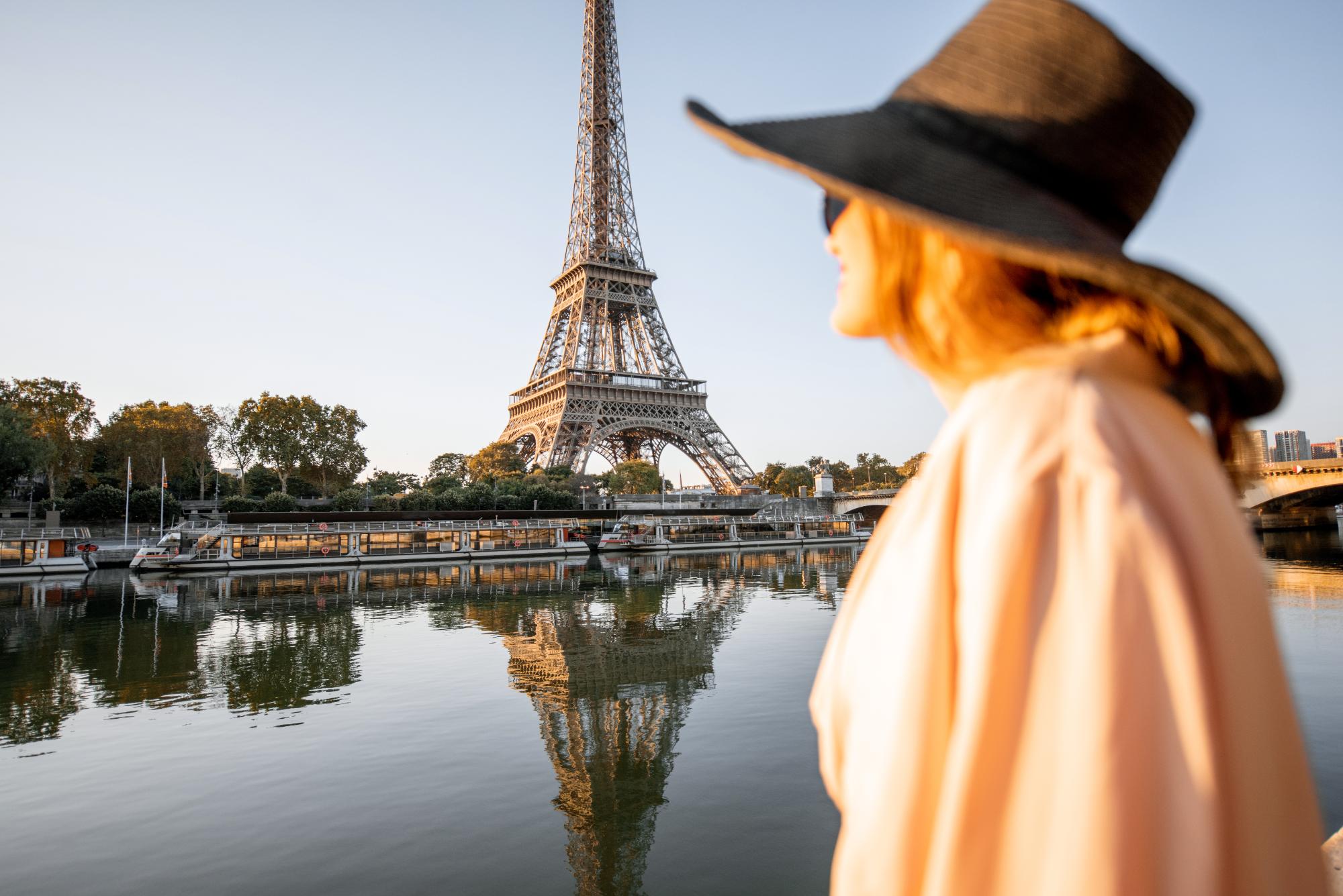 A woman in a large hat standing by the river, enjoying the view of the Eiffel Tower at sunset, with reflections in the water.