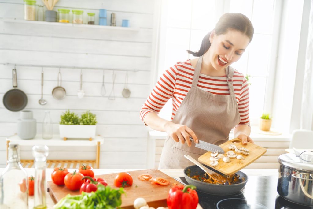 A woman in a striped shirt and apron is joyfully preparing a meal in a bright kitchen, chopping mushrooms and surrounded by fresh vegetables like tomatoes and lettuce.