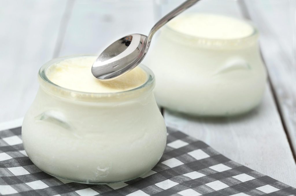 A close-up of a glass jar of creamy yogurt with a spoon resting on top, placed on a black and white checked napkin.