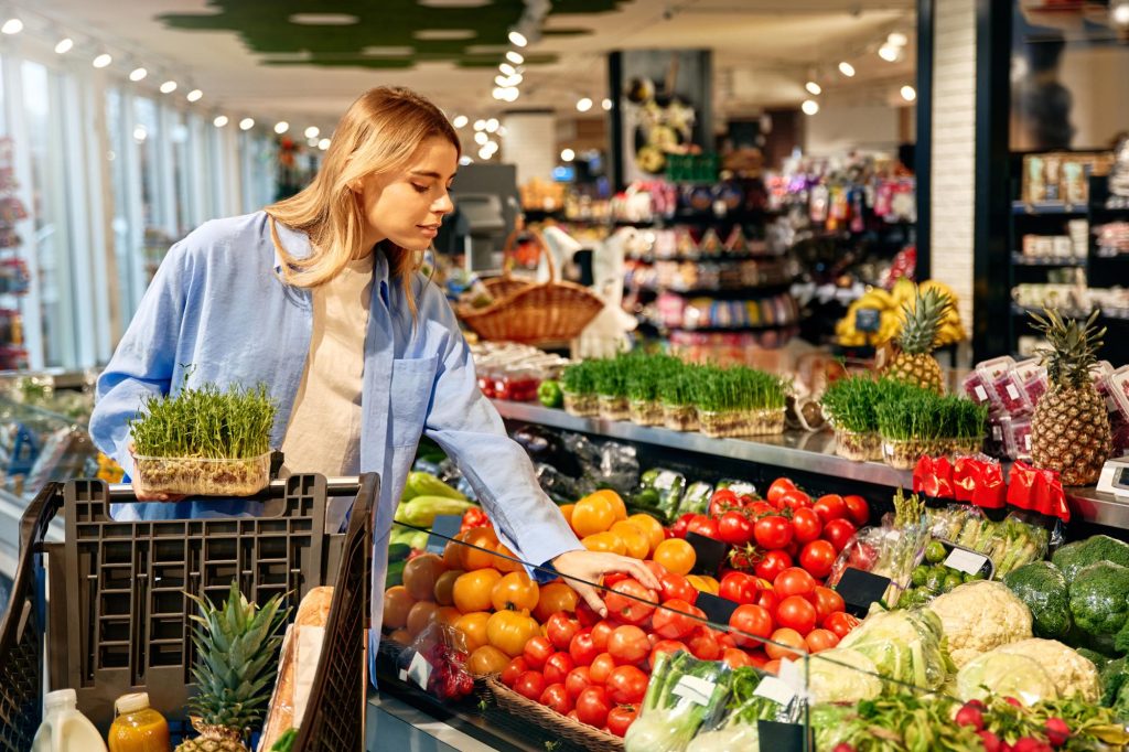 A young woman selecting fresh vegetables in an organic grocery store, holding a container of sprouts and examining tomatoes.