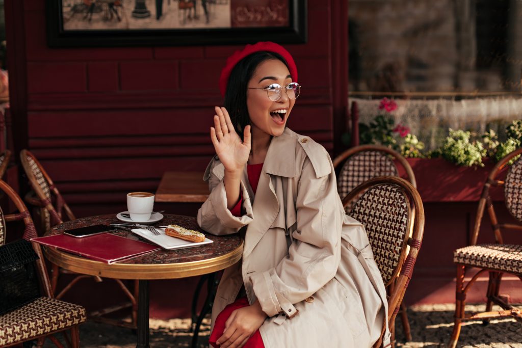 Happy brunette woman in trench coat, red dress and stylish beret waves hand in greeting, smiles widely and sits in street cafe.