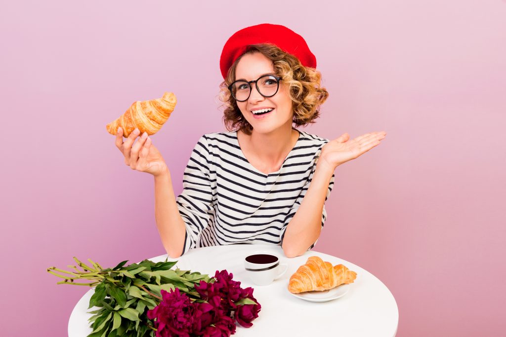 Happy traveling woman in France eating croissans with coffee , sits by the table over pink background.