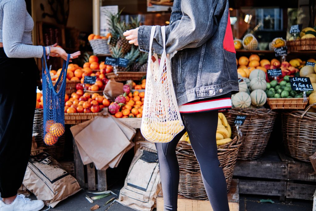 Crop unrecognizable female friends with reusable mesh bags choosing fresh fruits from stall at local market on urban street