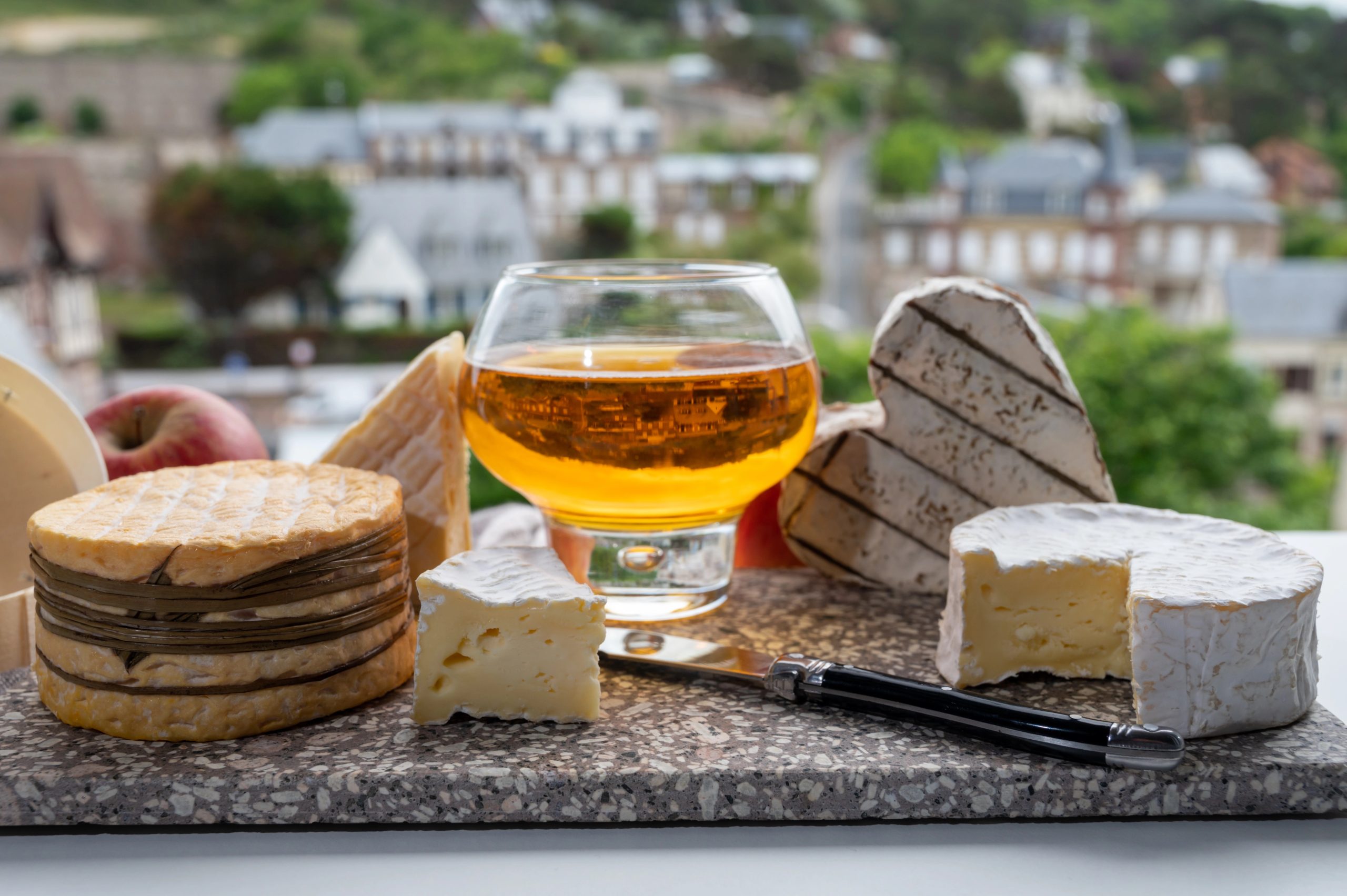 Cow cheeses of Normandy region - camembert, livarot, neufchatel, pont l'eveque and glass of apple cider drink with houses of Etretat village on background, Normandy, France